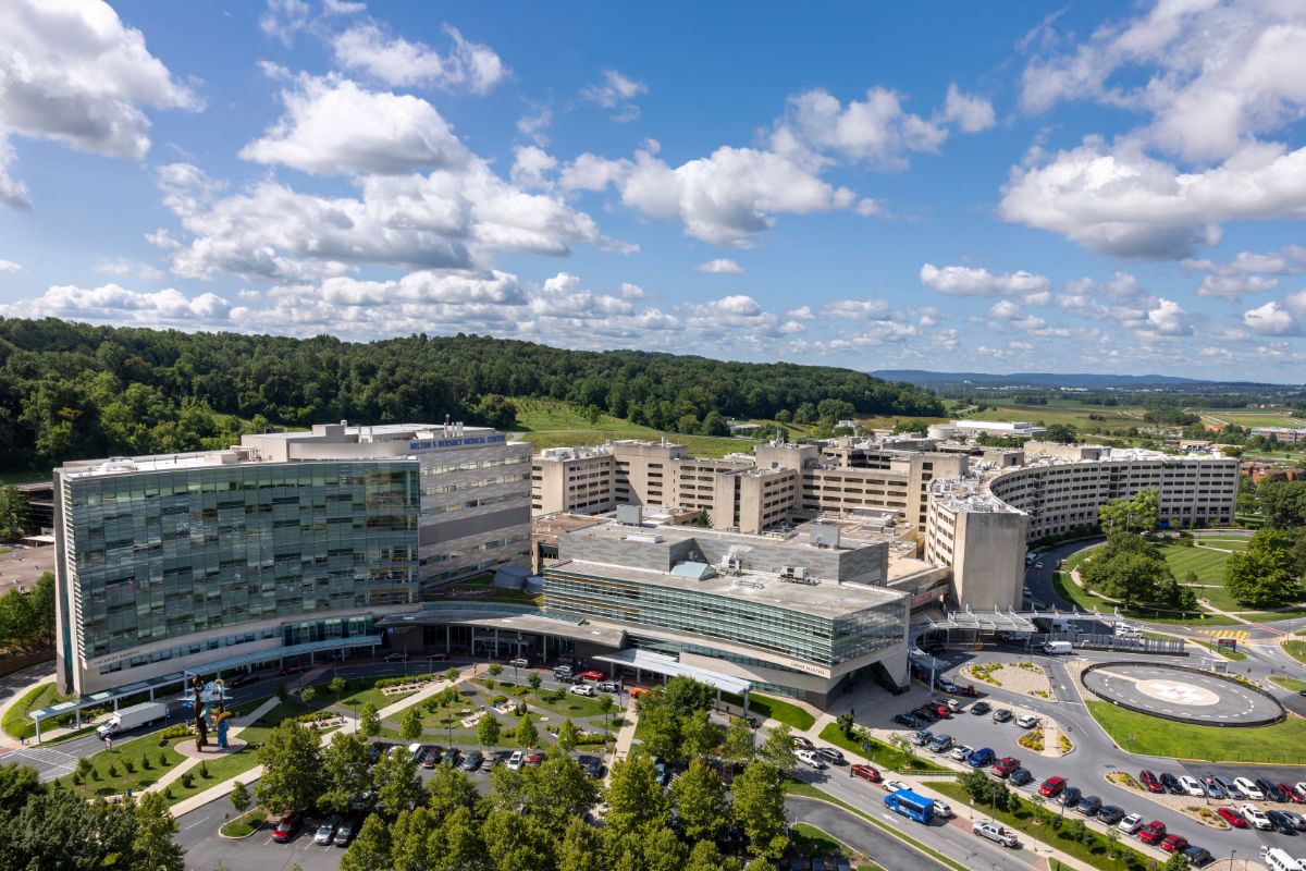 Aerial view of the Penn State Health Milton S. Hershey Medical Center campus with multiple modern buildings, parking areas, landscaped grounds, and a helipad, surrounded by wooded hills and farmland under a partly cloudy blue sky.