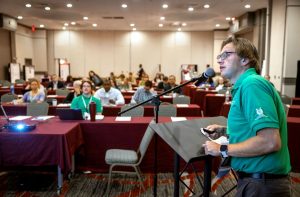 A man stands at a podium during a presentation he's giving to a large event space filled with people sitting at rows of tables.