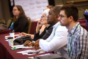 A close-up image of a row of people seated at a table, listening to a presentation.