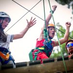 Three people stand atop a wooden platform, each wearing helmets and safety equipment related to ziplining. The child in the middle has his hands raised in the air.