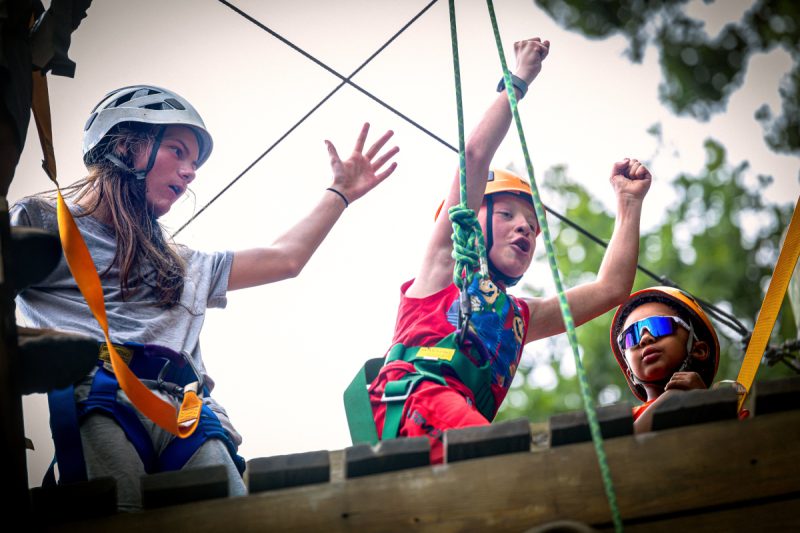 Three people stand atop a wooden platform, each wearing helmets and safety equipment related to ziplining. The child in the middle has his hands raised in the air.