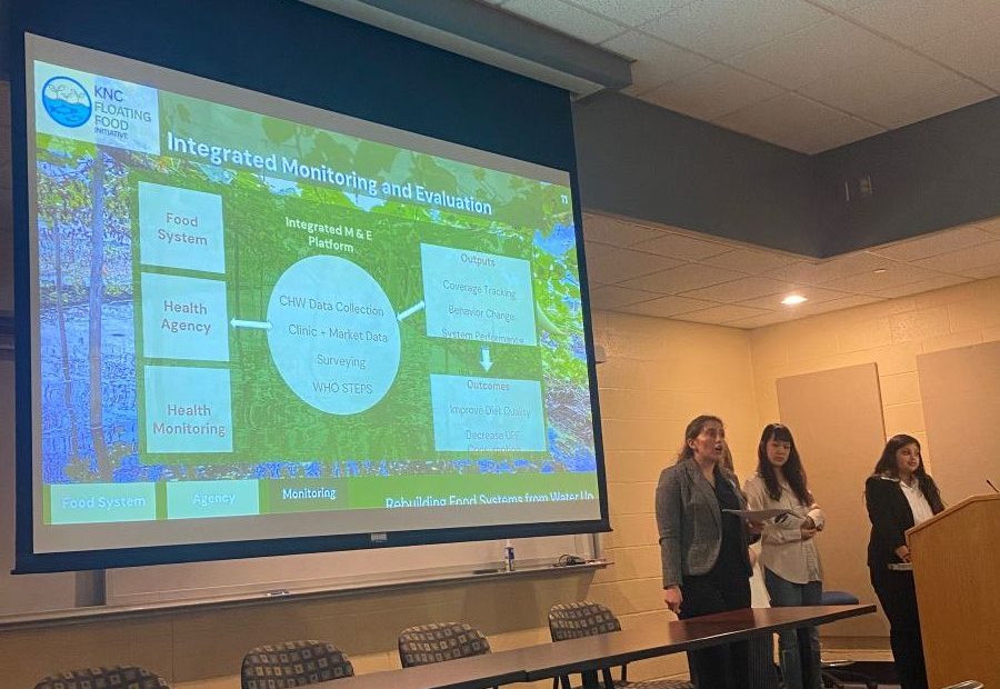 Students stand at a podium during a presentation that's displayed on a large projector screen behind them.