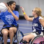 An individual in a wheelchair holds a basketball in the air and smiles. Another person to her right reaches out toward the ball.