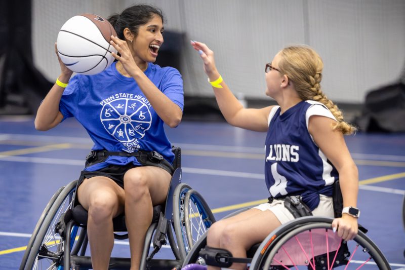 An individual in a wheelchair holds a basketball in the air and smiles. Another person to her right reaches out toward the ball.