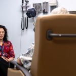 Dr. Sarah Kawasaki talks to a patient seated in an office exam chair. The patient is shown from behind. A blood pressure cuff and other medical instruments hang on the wall nearby.