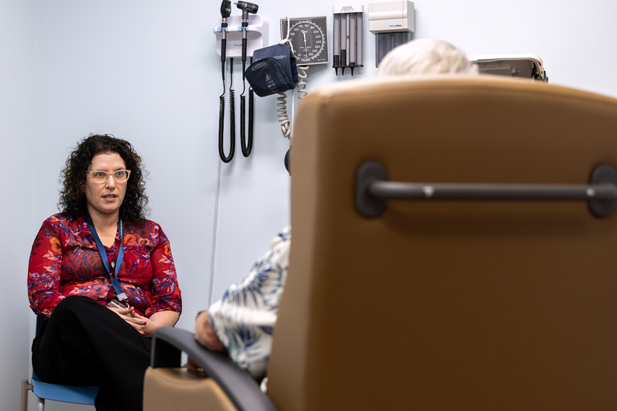 Dr. Sarah Kawasaki talks to a patient seated in an office exam chair. The patient is shown from behind. A blood pressure cuff and other medical instruments hang on the wall nearby.