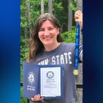 A woman holds a medal and certificate in front of trees.