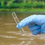 A gloved hand holds a beaker of water above a body of water