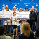 Group of Penn State Health physicians and staff hold oversized Hyundai Hope on Wheels checks totaling $500,000 for pediatric cancer research and care, with a child standing in front of the group.