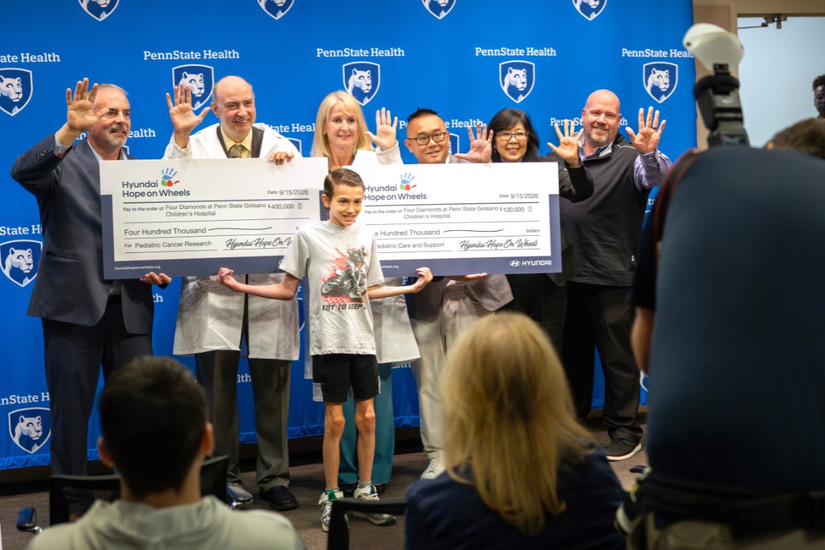 Group of Penn State Health physicians and staff hold oversized Hyundai Hope on Wheels checks totaling $500,000 for pediatric cancer research and care, with a child standing in front of the group.
