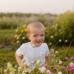 A young child wearing a white T-shirt sits among blooming wildflowers in an outdoor garden or flower field.
