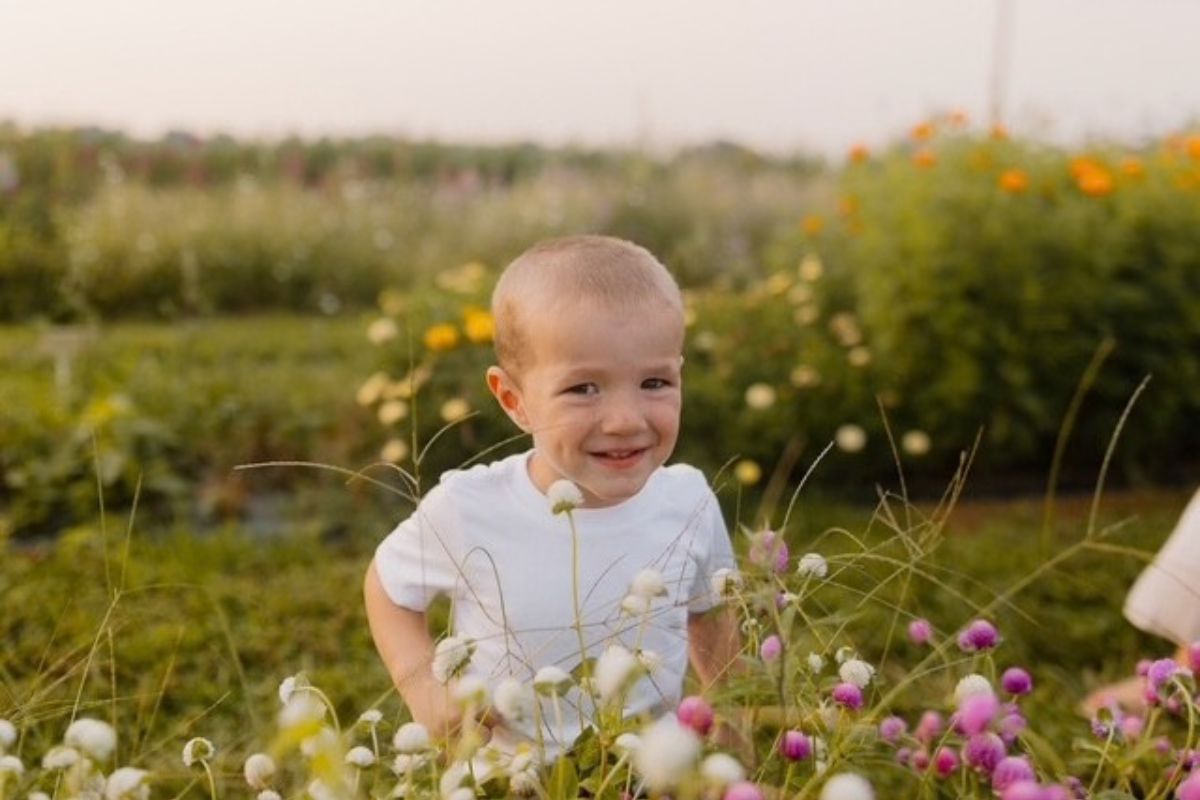 A young child wearing a white T-shirt sits among blooming wildflowers in an outdoor garden or flower field.