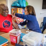 A man in a t-shirt smiles as a nurse gives him a flu shot in his left arm.