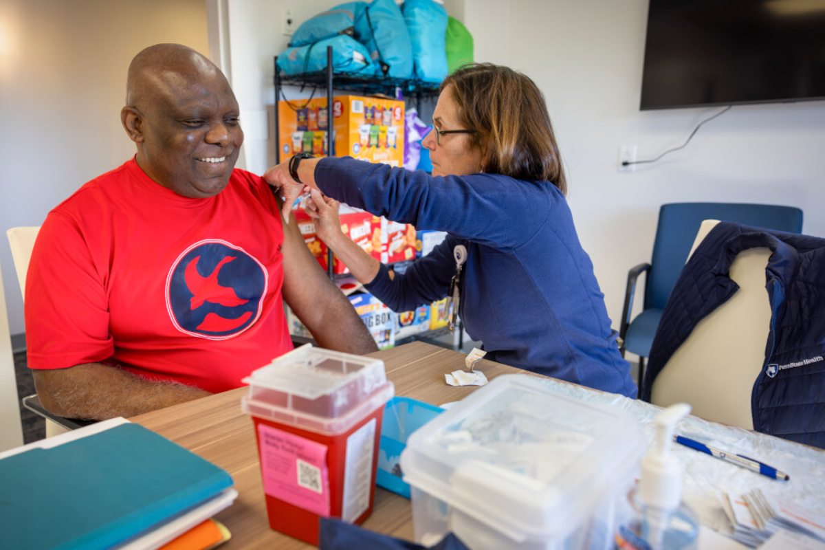 A man in a t-shirt smiles as a nurse gives him a flu shot in his left arm.