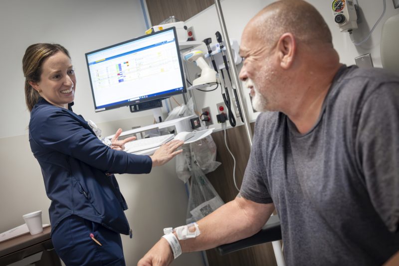 A patient sits in a chair in a clinical room, looking over at a nurse who stands at a computer terminal. She looks back, smiling.