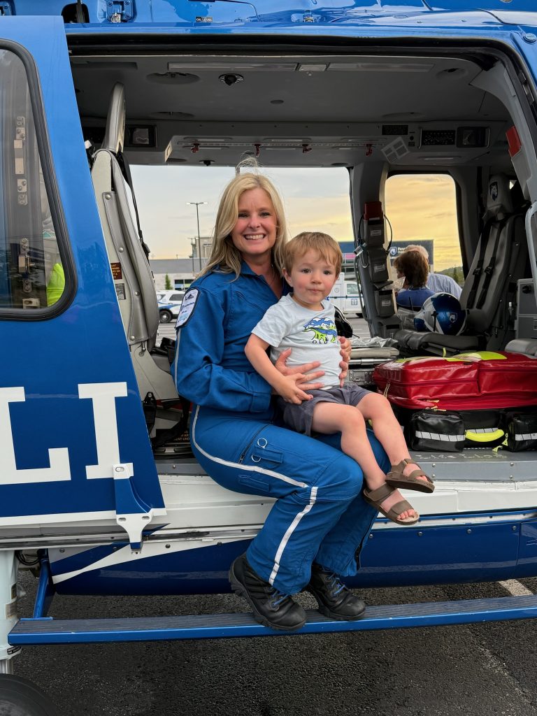A pediatric air medical transport team member in a blue flight suit sits at the open doorway of a medical helicopter while holding a young child on their lap.