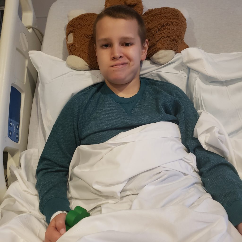 A child lies in a hospital bed covered with white blankets, holding a small green hand weight.