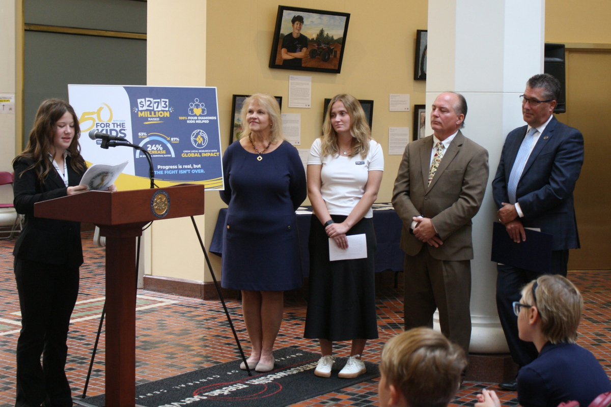 A teenage girl stands at a lectern delivering remarks. Four people stand nearby, looking on, as others – seated in front of her – listen to the comments.