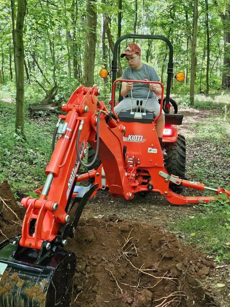 Taiven operates a compact tractor with a backhoe attachment on a wooded property, using the equipment to dig into the soil as part of outdoor landscaping work.