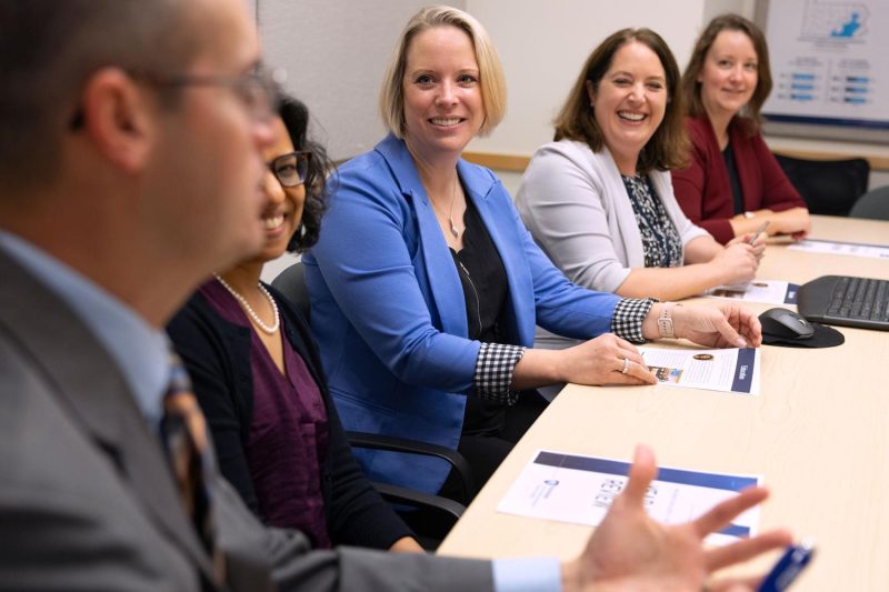 Penn State Clinical and Translational Science Institute leadership and scholars smile and listen attentively to a colleague speak during a meeting in a conference room.