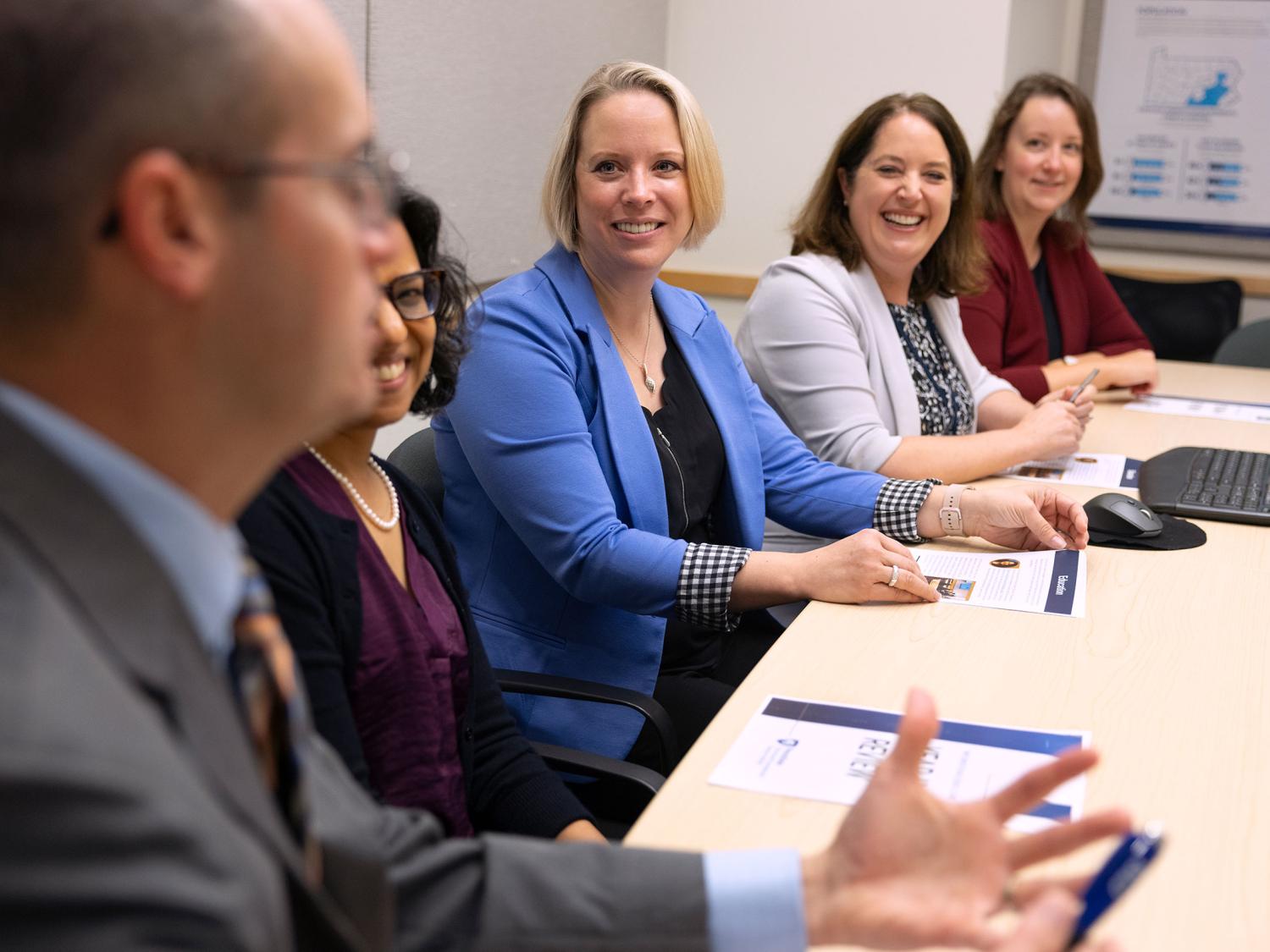 Penn State Clinical and Translational Science Institute leadership and scholars smile and listen attentively to a colleague speak during a meeting in a conference room.