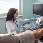 Healthcare provider in a medical exam room performing breast ultrasound exam on a woman.