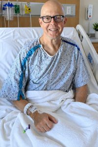 Stephen McNamara smiles from a hospital bed while wearing a hospital gown and holding an IV line.