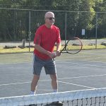 Stephen McNamara holds a tennis racket and prepares to return a shot on an outdoor tennis court, with a fence visible in the background.