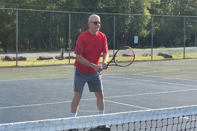 Stephen McNamara holds a tennis racket and prepares to return a shot on an outdoor tennis court, with a fence visible in the background.