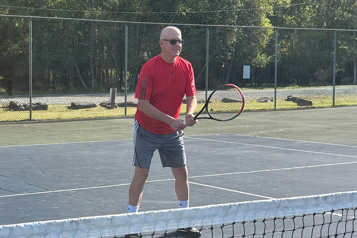 Stephen McNamara holds a tennis racket and prepares to return a shot on an outdoor tennis court, with a fence visible in the background.