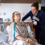 A nurse examines a hospitalized patient using a stethoscope while the patient sits upright in a hospital bed.