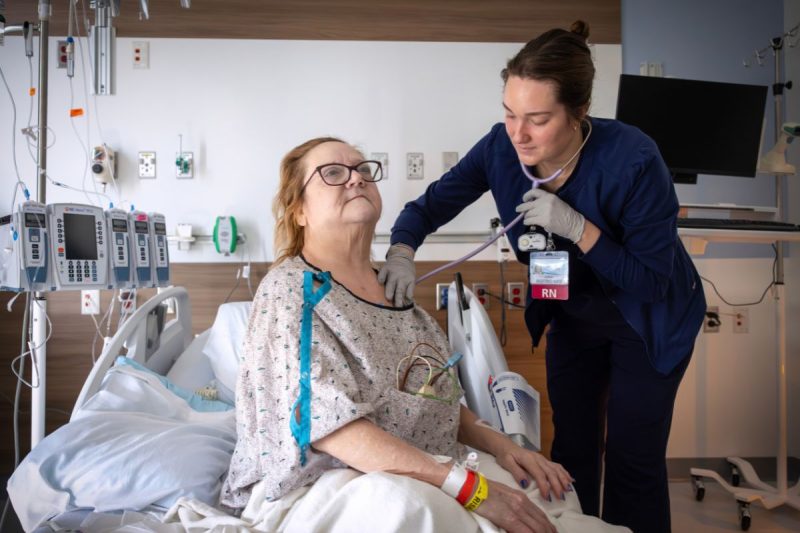 A nurse examines a hospitalized patient using a stethoscope while the patient sits upright in a hospital bed.