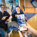 A man and a smiling young boy sit together in a hospital treatment area, with the man pointing toward the boy as another person photographs them with a smartphone.