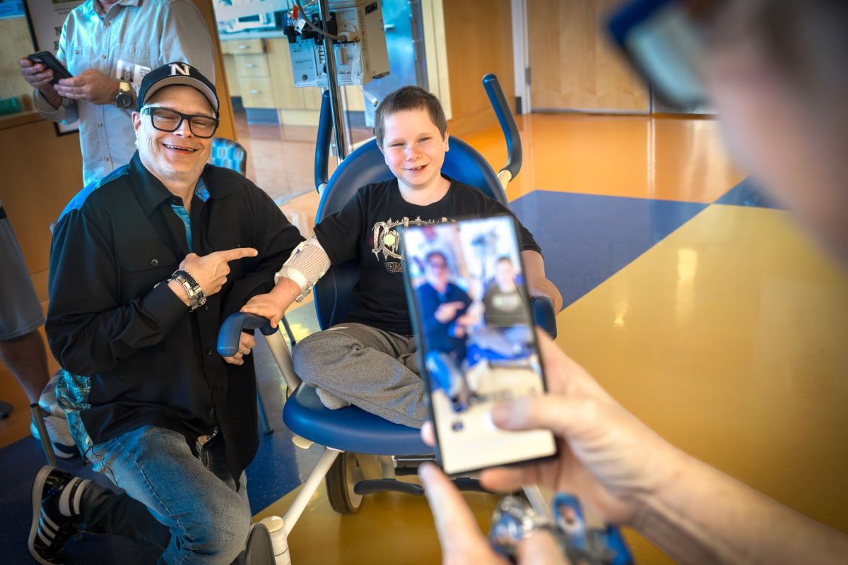 A man and a smiling young boy sit together in a hospital treatment area, with the man pointing toward the boy as another person photographs them with a smartphone.