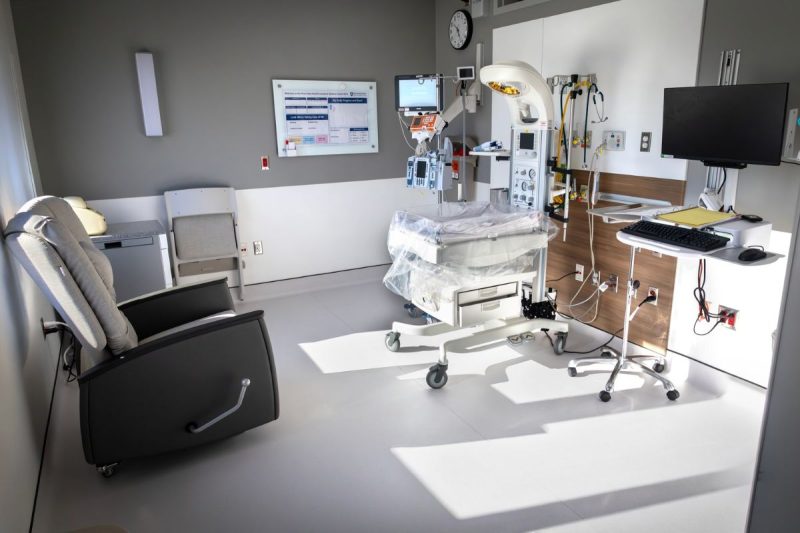 Bright, private neonatal intensive care unit (NICU) room at Penn State Health Lancaster Medical Center featuring an infant warmer with monitoring equipment, IV pumps, overhead exam light, bedside computer workstation, and a reclining chair for parents.