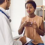A patient sitting in a medical exam room, speaking earnestly with a doctor who is taking notes on a clipboard.
