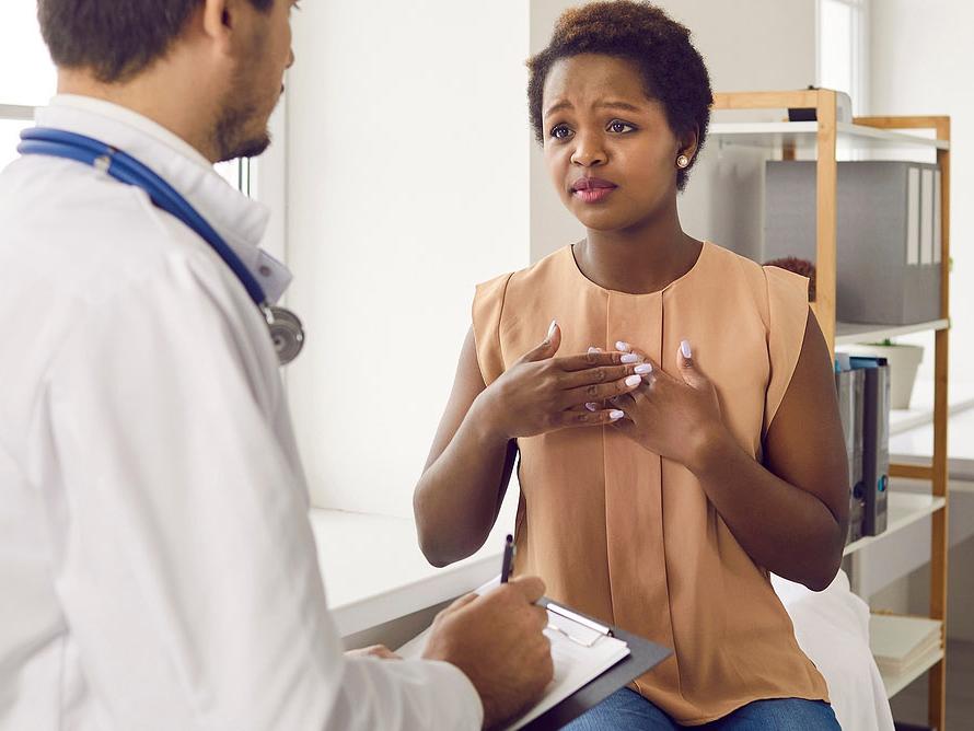 A patient sitting in a medical exam room, speaking earnestly with a doctor who is taking notes on a clipboard.