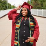 A smiling pregnant woman poses outdoors in a red graduation cap and gown, wearing a black dress and a colorful stole reading “Black Girl Magic” as she cradles her belly.