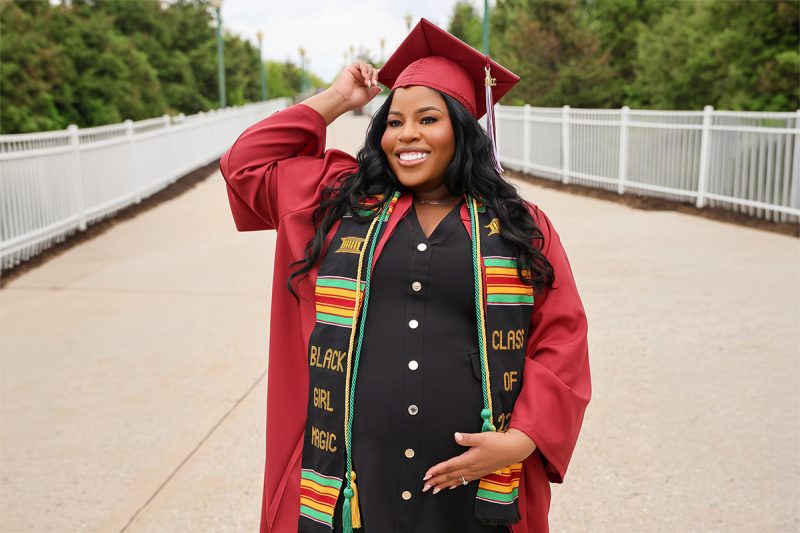 A smiling pregnant woman poses outdoors in a red graduation cap and gown, wearing a black dress and a colorful stole reading “Black Girl Magic” as she cradles her belly.