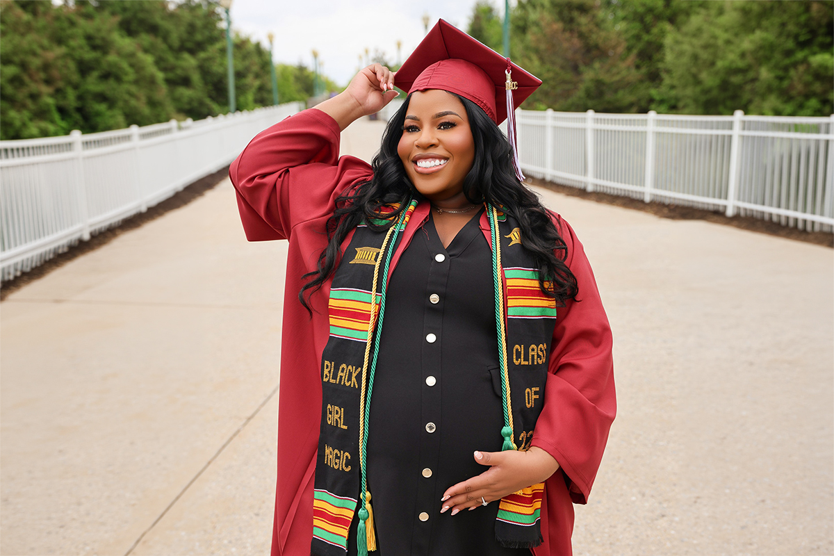 A smiling pregnant woman poses outdoors in a red graduation cap and gown, wearing a black dress and a colorful stole reading “Black Girl Magic” as she cradles her belly.